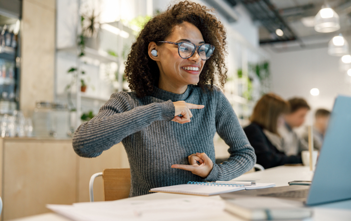 Una mujer habla en lengua de señas frente a una computadora, usa lentes y tiene un auricular en su oreja derecha, está sentada en un escritorio. En el fondo, desenfocadas, hay tres personas también en un escritorio