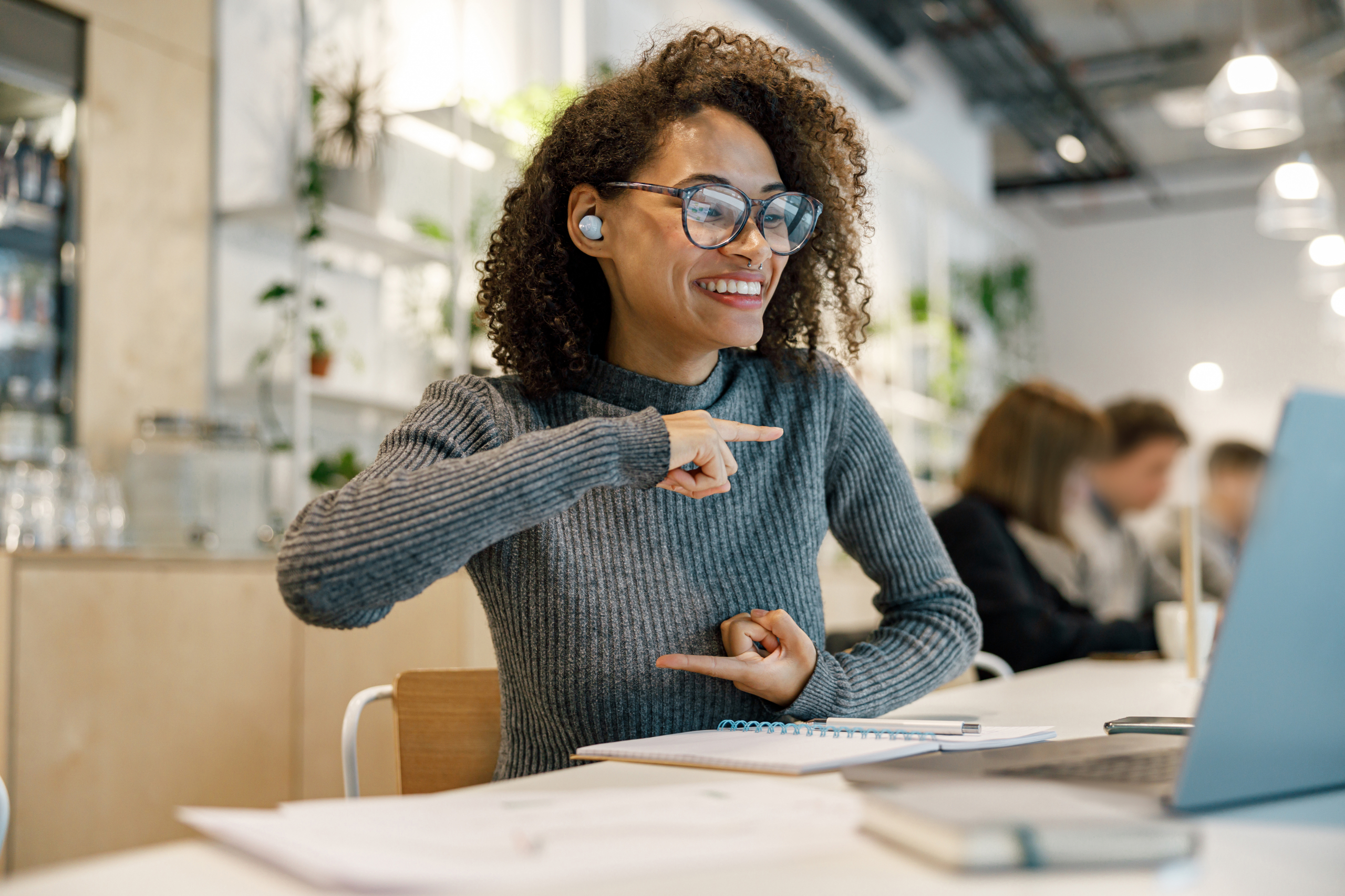 Una mujer habla en lengua de señas frente a una computadora, usa lentes y tiene un auricular en su oreja derecha, está sentada en un escritorio. En el fondo, desenfocadas, hay tres personas también en un escritorio